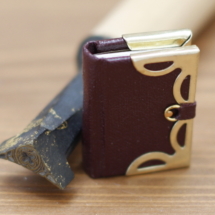 A miniature book with brass corners and clasp, displayed with a book finishing tool larger than the book.